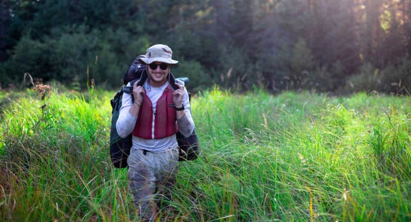 A person wearing a lifejacket carries gear through tall green grass. 